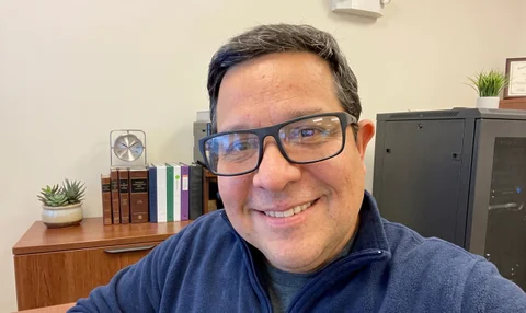 Man in glasses smiling in an office with books and plants.